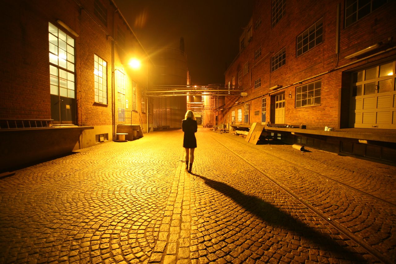 A woman walks alone down a cobblestone alley illuminated by streetlights.
